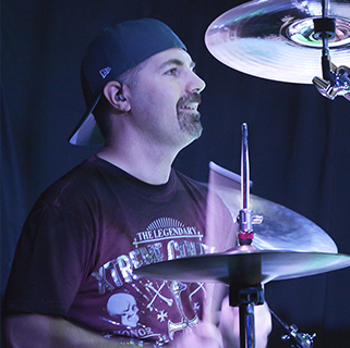A drummer playing on stage, wearing a backward baseball cap and a graphic T-shirt. The drum set and cymbals are partially visible, with stage lighting casting a blue hue on the scene.