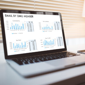 A laptop screen displays charts and graphs titled "Email by DMU Member," with categories for Cybersecurity, Tech Architect, Tech Exec, and User Cruise. Each section shows open and total click rates, with bar graphs and percentage data.