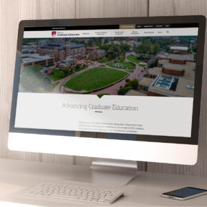 A computer screen displays a university's website. The homepage features an aerial view of a campus with green spaces and buildings, along with the text "Advancing Graduate Education" and information about graduate programs.