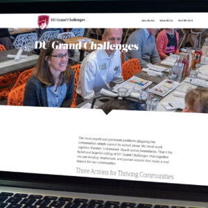 A group of people sits around tables covered with papers and glasses, engaged in discussion. In the background, text reads "DU Grand Challenges." The website header includes navigation links like "Who We Are" and "What We Do.