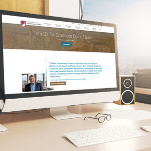 A desktop computer displays a university website advertising a graduate open house event. A pair of glasses and speakers are placed next to the monitor. The screen shows an image of a man and a quote about addressing global challenges.
