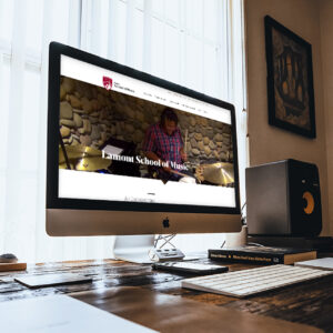A modern desk setup with an iMac displaying the "Lamont School of Music" website. The workspace includes a speaker, keyboard, mouse, and stacked books, with a sunlit window and framed pictures in the background.