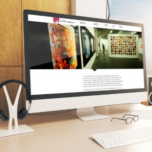 A computer screen displays a website featuring an art and art history school with a gallery exhibit. The desk has a pair of headphones on a stand and a smartphone with glasses placed nearby. A sunny window casts natural light on the setup.