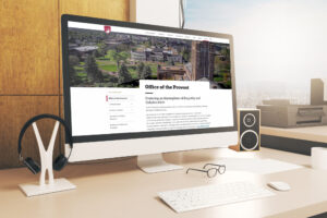 A computer on a desk displays the "Office of the Provost" webpage. The room has a view of a cityscape, and the desk holds a keyboard, mouse, headphones, and speakers. Sunlight illuminates the scene.
