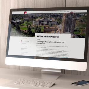 A computer monitor displays a webpage titled "Office of the Provost" featuring an aerial view of a university campus. A keyboard and mouse sit on the wooden desk below the monitor.
