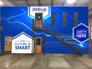 A blue wall inside a building features a staircase with black railings. Signs on the wall read, "The Future is Smart" and "Your Journey to Connected Power Begins Here," accompanied by white geometric designs. There are three doors and three windows.