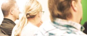 A group of three people is sitting and facing forward, possibly listening to a presentation. The focus is on the back of their heads and shoulders. One person has a ponytail, and the others appear to have short hair.