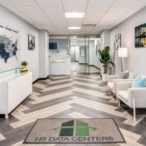 Modern office lobby with a chevron-patterned floor, white cushioned chairs, and a welcome mat labeled "H5 Data Centers." Walls display maps and framed pictures, and a potted plant adds greenery. Glass doors lead to a hallway in the background.