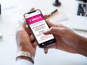 A person holding a smartphone displaying a website titled "T: Minus 10" with sections about what's new, social media, and events. The phone is held in one hand while the other hand scrolls the screen. A desk with office items is in the background.