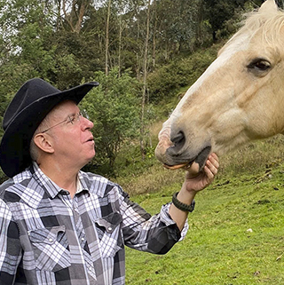 A person in a plaid shirt and cowboy hat is feeding a light-colored horse in a grassy, wooded area. The person looks up at the horse with a soft expression, while the horse gently takes food from their hand.