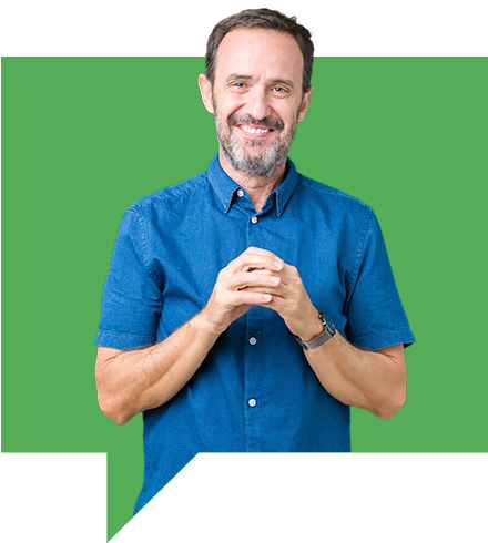 A man with a beard smiles while standing against a green background. He is wearing a short-sleeved blue button-up shirt and holding his hands together in front.