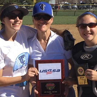 Three women on a softball field pose for a photo. The woman in the middle holds a USSSA plaque, and the woman on the right holds an MVP award. They are smiling and wearing athletic gear.