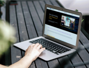 Person using a laptop on a wooden table, viewing a website with video content and a banner saying "No Gimmicks Just Plenty of Fiber." The person's hand is on the keyboard, and there are blurred green plants in the background.