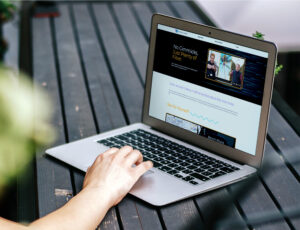 A person uses a laptop on a dark wooden table, browsing a website with a banner that reads "No Gimmicks, Just Plenty of Fiber." The screen shows images and text. Greenery is blurred in the foreground.