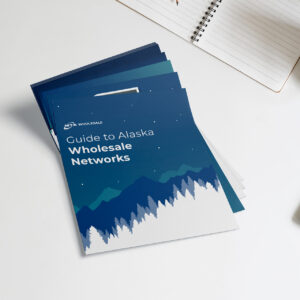 Stack of brochures titled "Guide to Alaska Wholesale Networks" with a design featuring mountains and trees against a blue background. A spiral notebook is partially visible in the top right corner on a white surface.