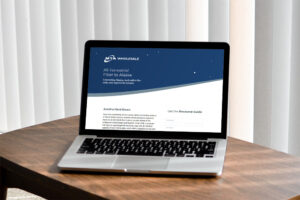 A laptop on a wooden table displays a website for MTA Wholesale, featuring information about all-terrestrial fiber to Alaska. The screen shows text under headings "Avoid a Hard Down" and "Get the Resource Guide." Vertical blinds are in the background.