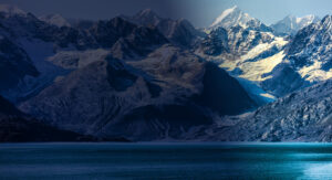 A dramatic mountain range, partially in shadow, with jagged snow-capped peaks. The lower portion is illuminated by sunlight, creating a stark contrast. A serene blue body of water lies in the foreground.