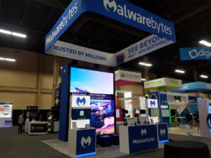 A technology exhibition booth for Malwarebytes, showcasing large screens with digital displays. The booth has banners saying "Trusted by Millions" and "See Beyond Next-Gen Antivirus," with tables displaying promotional materials.