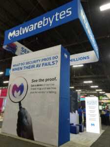 Image of a Malwarebytes trade show booth featuring large hanging banners. A central display reads "What do security pros do when their AV fails?" with the company's logo on a magnifying glass. There are also seating areas and informational graphics.