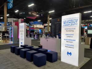A convention booth with a large banner displaying "EDR Capabilities Without Complexity," highlighting features like continuous monitoring, endpoint isolation, ransomware rollback, and complete remediation. Blue seating cubes are arranged in front.