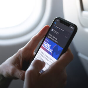 A person holding a smartphone displaying an email about a 2019 Educause event. The screen shows blue and white text with a partial view of an airplane window in the background, suggesting they are on a flight.