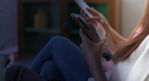 A person is sitting comfortably, using a smartphone. They have crossed legs and are wearing jeans. The room is softly lit, creating a relaxed atmosphere. Only the person's hands and knees are visible.