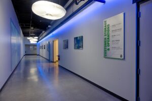 A modern hallway with polished floors and blue accent lighting. Large white fixtures hang from the ceiling. Various framed pictures and a green sign labeled "CUSTOMER STORAGE & OPERATIONS" adorn the walls.