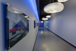 A modern hallway with a polished concrete floor, blue accent lighting on the ceiling, and large, circular light fixtures. Framed images line the walls, featuring a glass-encased structure and the words "LONG LIVE YOU.