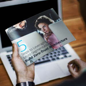 A hand holding a brochure titled "5 Steps to Leveraging Micro-Segmentation to Build Zero-Trust Architecture." In the background, a laptop is partially visible on a wooden table.