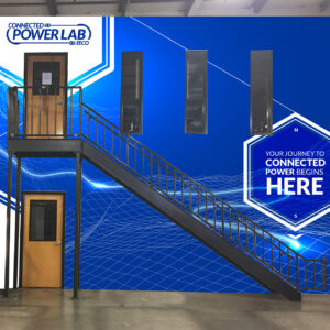 A blue wall with "Connected PowerLab" and "Your journey to connected power begins here" text. It features geometric patterns and a staircase leading to an elevated platform with a door and four narrow windows.