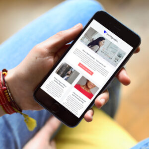 A person holds a smartphone displaying a newsletter from a health-related website. The screen shows images and text sections, with a prominent "Read More" button. The phone is held in a hand wearing a bracelet.