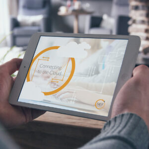 A person holds a tablet displaying a screen with the text "Connecting to the Cloud" and cloud graphics. The background is blurred, showing a cozy room with a chair and table.