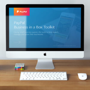 A desktop computer displaying the "PayPal Business in a Box Toolkit" on the screen. The desk has a keyboard, a mouse, and a set of colorful markers.