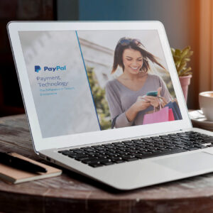 A laptop on a table displays a PayPal presentation about payment technology. The screen shows a smiling woman holding a phone and shopping bags. A notebook and pen are nearby.