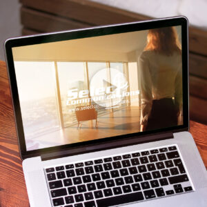 Open laptop displaying a website with the logo "Select Communications" and a URL. The screen shows a person standing in a room with large windows and a city view.