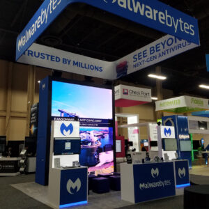 A booth at a tech convention showcases Malwarebytes, featuring a large screen displaying security software information, branded kiosks, and promotional banners. Nearby booths are visible in the background.