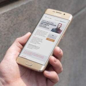A hand holding a Samsung smartphone displaying a webpage about cybersecurity solutions. The page includes text about security options and a call to action button. The background is a blurred stone surface.