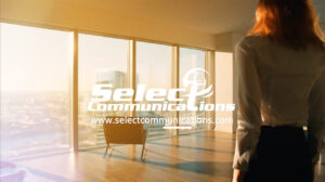 A person stands in front of large windows with a view of city buildings under a bright sky. The floor is wooden, and there is a chair facing the windows. The text reads "Select Communications" with their website address.