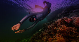 A snorkeler in a wetsuit swims underwater near a vibrant coral reef. Sunlight filters through the water, creating a purple hue. The scene is serene, showcasing diverse marine life and colorful corals.