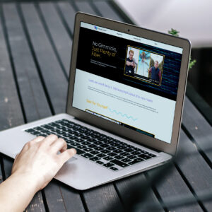A person using a laptop on a wooden table. The screen displays a website with a banner at the top saying, "No Gimmicks, Just Plenty of Fiber," featuring a video with two people.