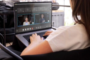 A person sits with a laptop on their lap, participating in a video conference. The laptop screen shows two speakers, a woman and a man, in online meeting windows.