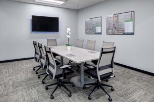 A modern conference room with a long table surrounded by eight office chairs. A potted plant sits on the table. A flat-screen TV is mounted on the wall, and two detailed maps are displayed beside it. The room has a neutral color scheme.