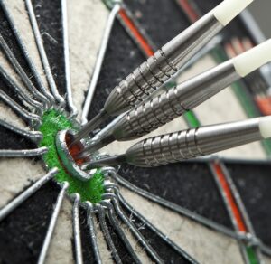Close-up of three darts perfectly hitting the bullseye on a dartboard. The darts are metallic with white shafts, and the board features green, black, and red colors.