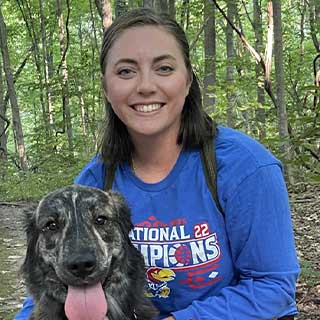 A woman in a blue shirt smiling while kneeling next to a happy dog with its tongue out. They are surrounded by a forest with green trees.
