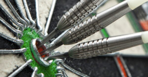 Three silver darts are embedded in the bullseye of a dartboard. The focus is close-up, highlighting the precision of the throw, with the surrounding dartboard colors visible.