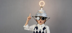 A child wearing aviator goggles and a vest points upward with one finger. They have a metal colander helmet with a glowing light bulb on top, set against a plain gray background, expressing an idea or inspiration.