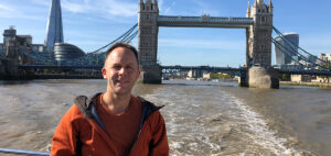 A person stands on a boat with Tower Bridge and the Shard in the background. The sky is clear and the river reflects the sunlight. The person is wearing an orange jacket and smiling at the camera.