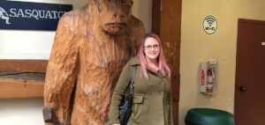 A woman with pink hair stands smiling in front of a large wooden Sasquatch statue. She is wearing a green coat and holding a black bag. There is a sign above her and a WiFi symbol on the wall.