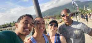 Four people smile for a selfie at a sunny, tropical location. Mountains and palm trees are in the background, along with people walking nearby. The atmosphere is cheerful and relaxed.
