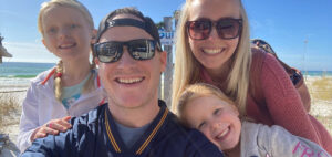 A family of four, including two young children, is smiling and posing for a selfie on a sunny beach. The ocean is visible in the background, and they are dressed in casual, warm-weather clothing.